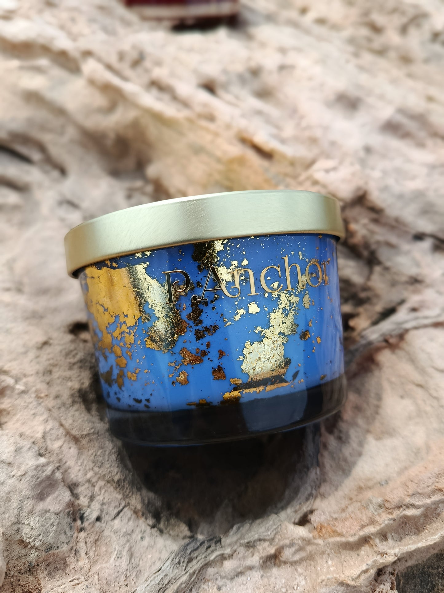 Luxury blue P.Anchor candle with gold flakes and metallic lid, placed on natural rock surface.
