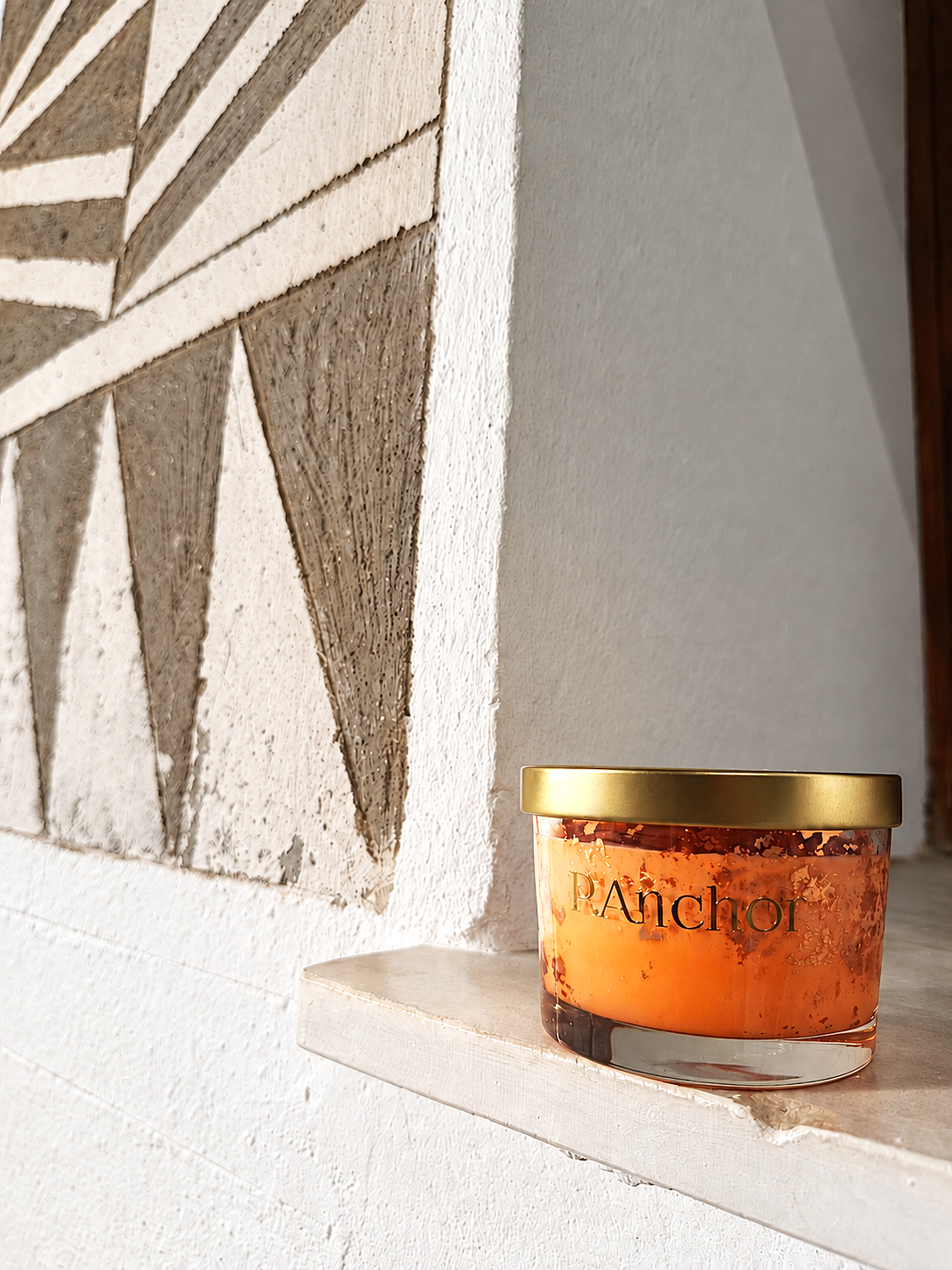 A glass jar candle with amber-orange wax and a gold lid sits on a stone ledge next to a textured white and gray geometric wall.