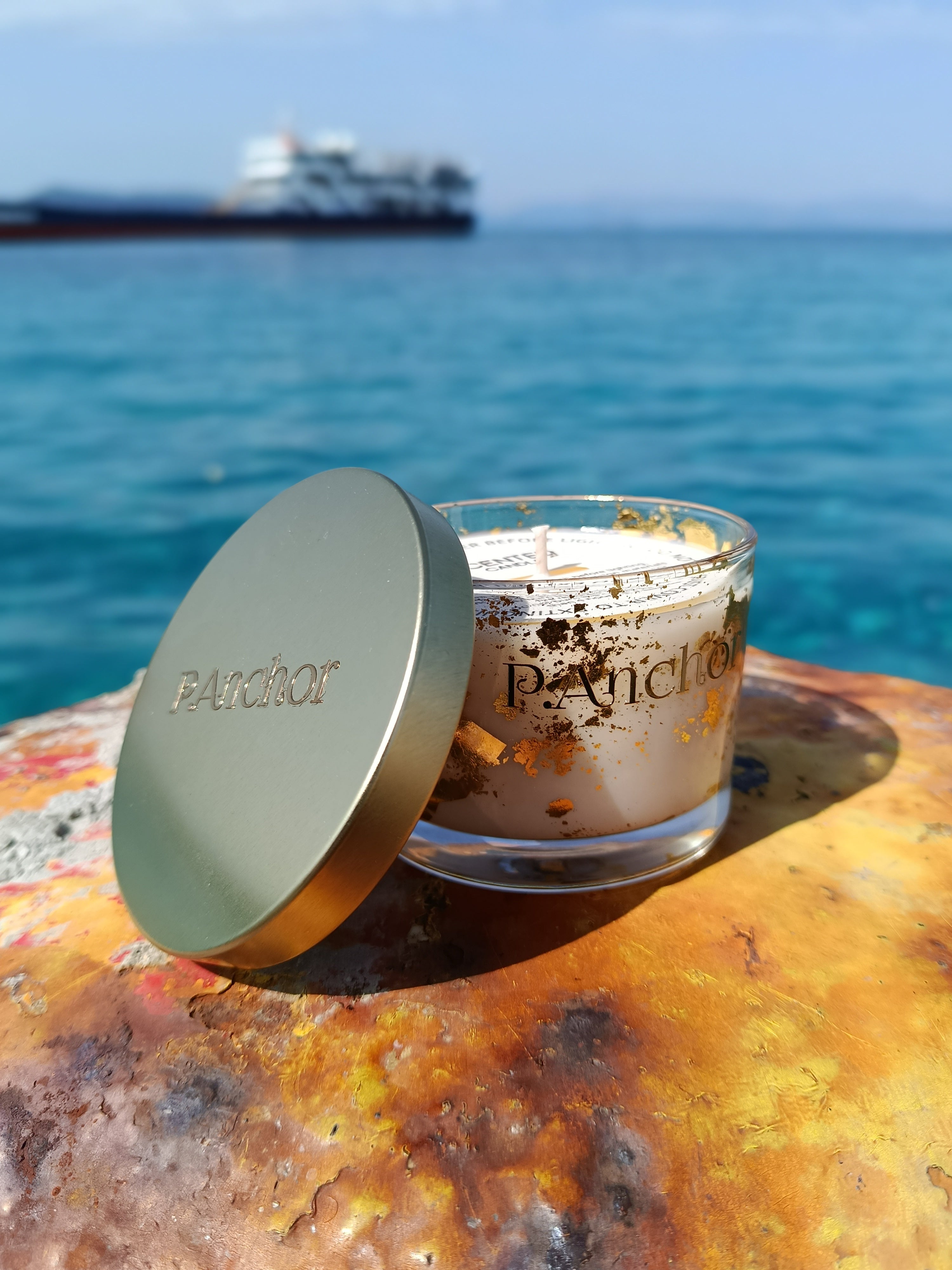 P.Anchor luxury candle with gold lid and gold flakes, placed by the sea with a ship in the background