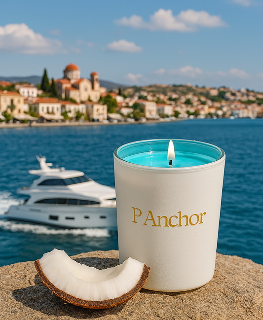 P.Anchor coconut and cashmere scented candle with aqua blue wax and gold logo, styled with coconut slice by the sea with yacht and Greek village background