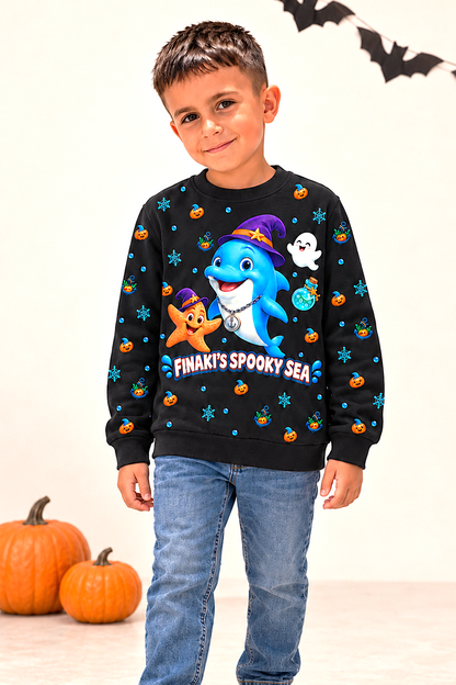 A young boy wearing a black long-sleeve crewneck sweatshirt with a colorful cartoon graphic of a shark in a witch hat and the text 'FINAKI'S SPOOKY SEA'. The sweatshirt features small orange pumpkins, blue stars, and white ghosts.