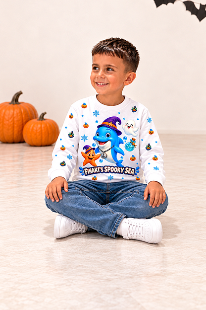 A young boy sits cross-legged on a light beige floor wearing a white long-sleeve Halloween sweatshirt with a colorful cartoon shark graphic and the text 'FINKY'S SPOOKY SEA'. The background features a warm off-white wall with a pumpkin decoration on the left.