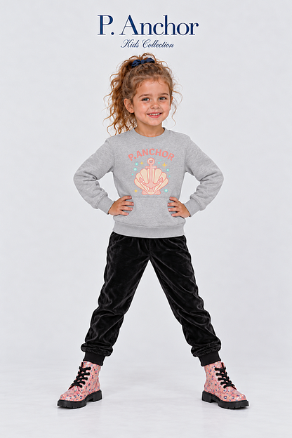 A young girl stands in a studio wearing a light gray sweatshirt with a pink shell graphic and the word 'PANCHOR,' black velvet pants, and pink patterned boots. The brand name 'P.