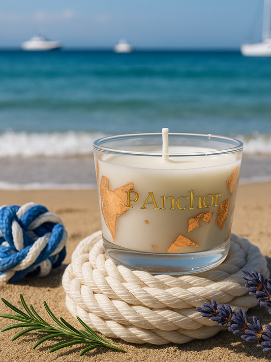 A luxury P. ANCHOR candle in a clear glass jar with gold foil accents, placed on coiled white nautical rope on a sandy beach. The background shows the sea with yachts and a clear blue sky, surrounded by sprigs of rosemary and lavender, and a decorative navy-blue rope.