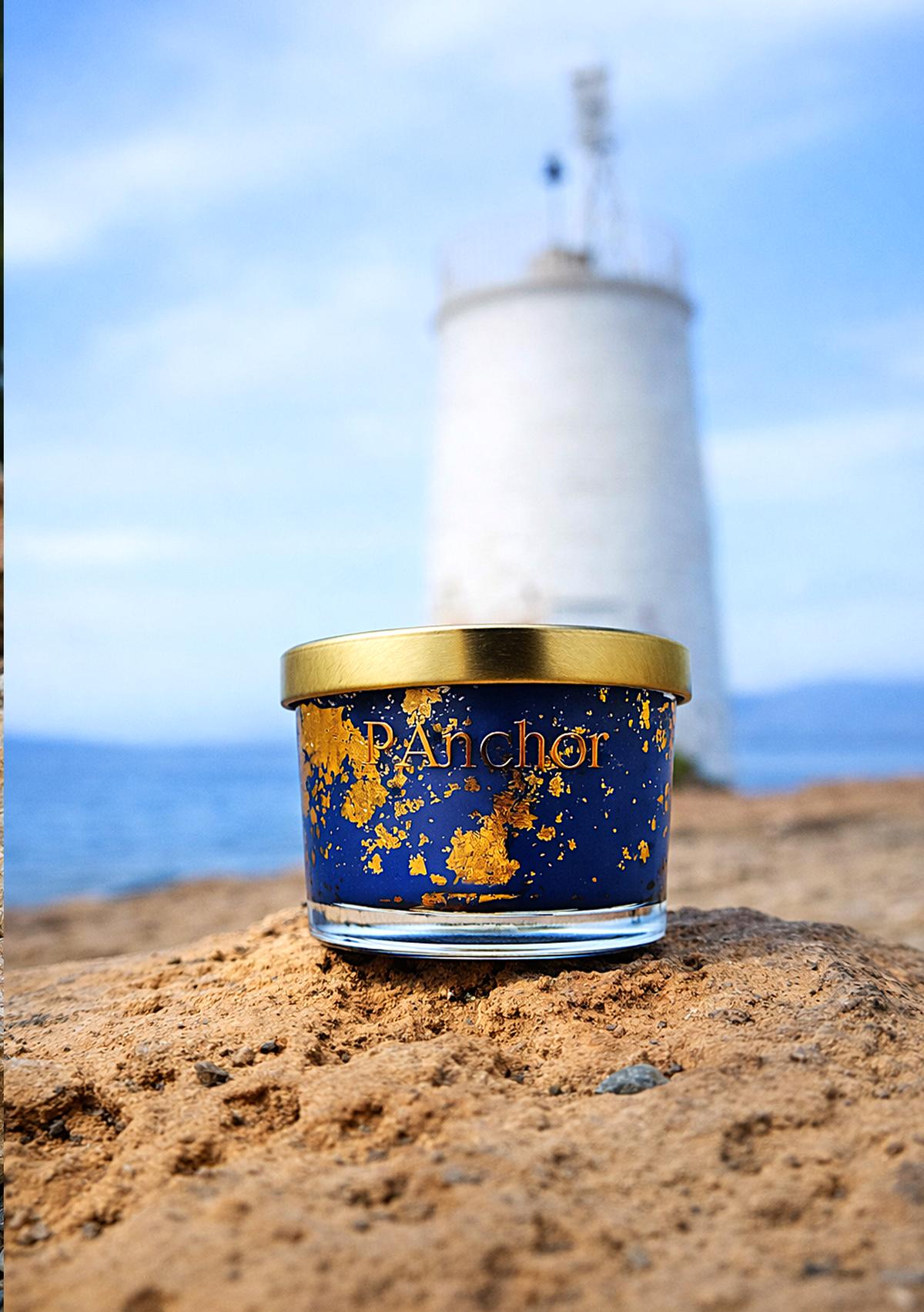 A small glass jar candle with a gold lid and a deep blue label featuring gold leaf splatter and the brand name 'Pancher' sits on a sandy beach with a blurred lighthouse and ocean in the background.
