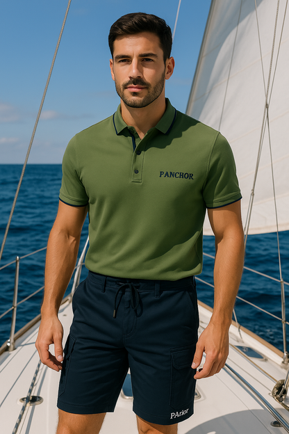 Man standing on a sailboat wearing a green P.Anchor polo shirt and navy blue cargo shorts with "PAnchor" logo, with the sea and sail in the background.