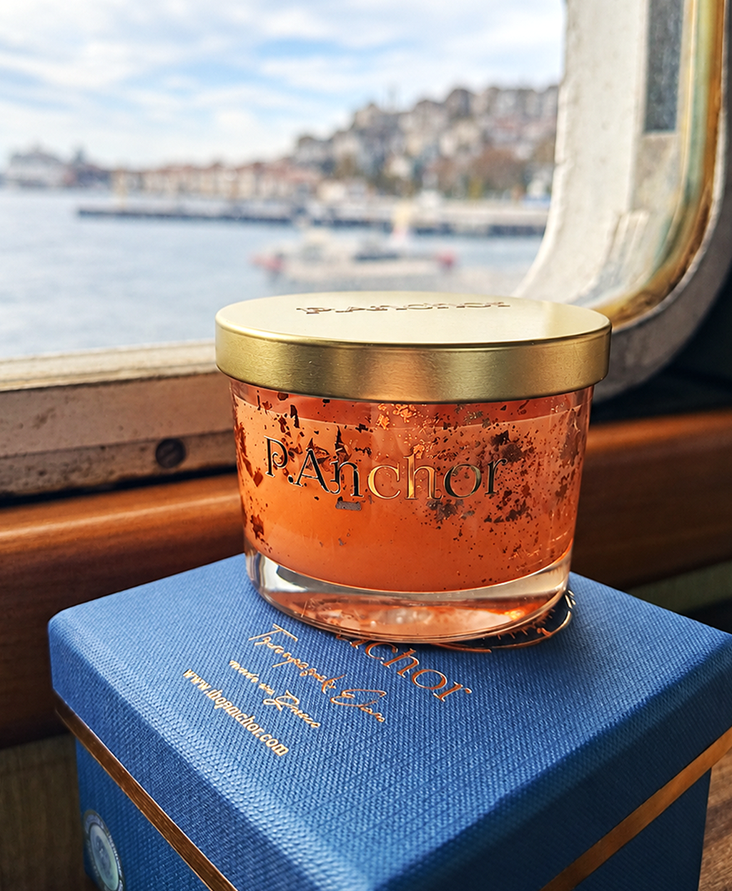 A jar of Panachot body butter with orange body butter and gold foil flakes, resting on its blue branded box on a train window seat with a blurred Istanbul harbor background.