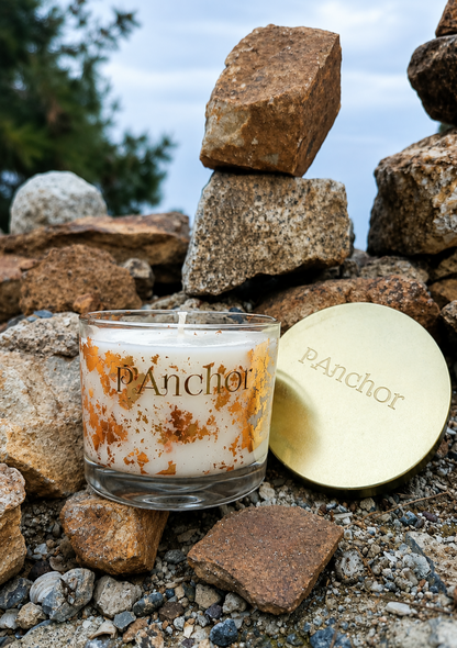 A glass jar candle with white wax and orange-brown speckled detailing sits outdoors among stacked rocks and gravel, with its round gold lid placed beside it. The lid and candle are branded 'pAnchor'.