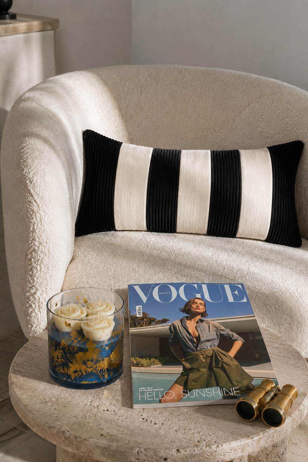 Decorative pillow on a white armchair with a 'Vogue' magazine and candles on a stone table.