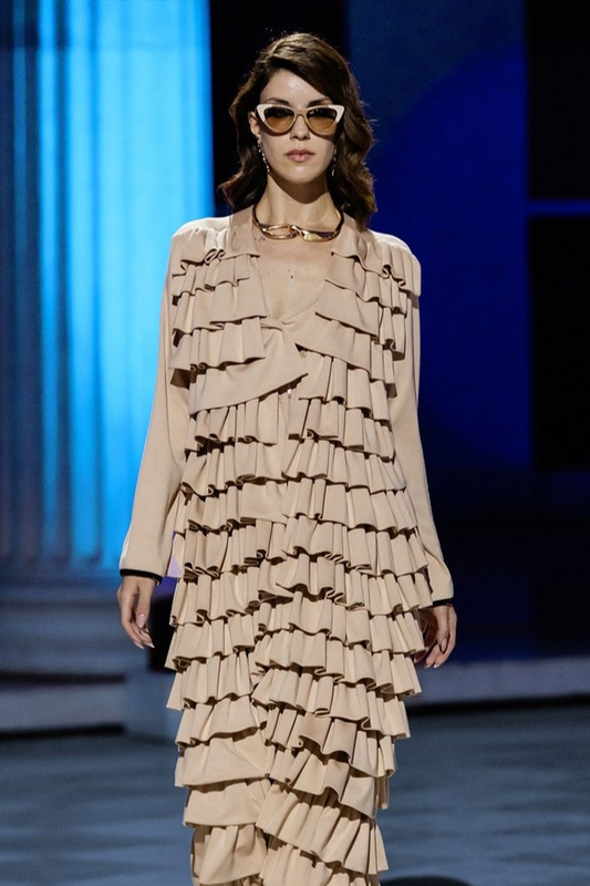 Model wearing a beige ruffled dress on a runway with a blue curtain background